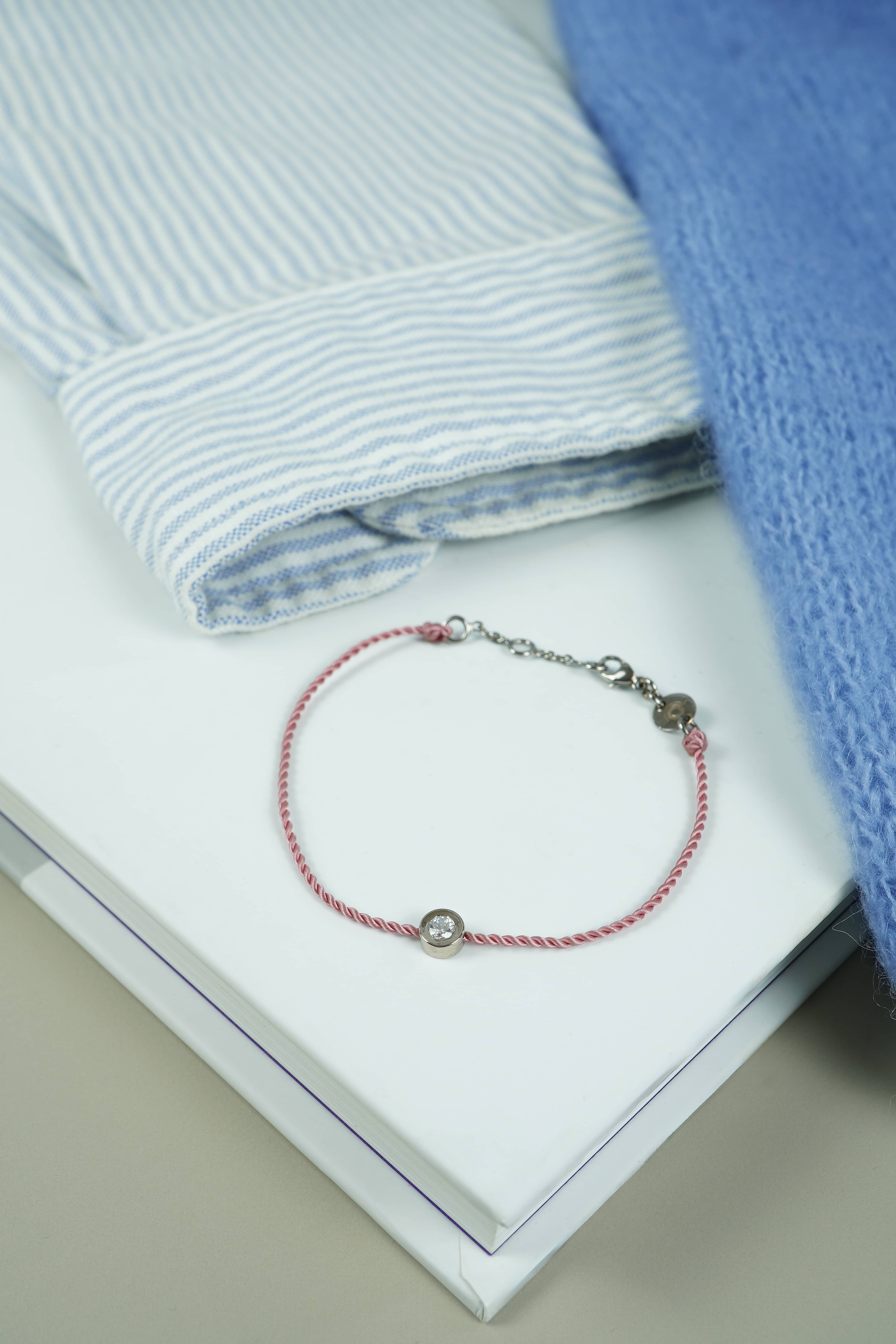 Bracelet ONE cordon rose foncé et or blanc, posé près d'une chemise rayée et d'un pull bleu