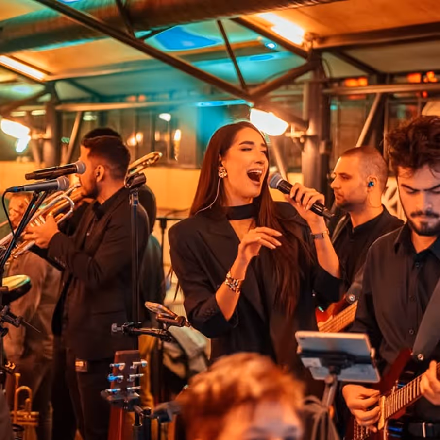 Muzičari izvode uživo, pevačica drži mikrofon, a drugi sviraju gitare i trubu.