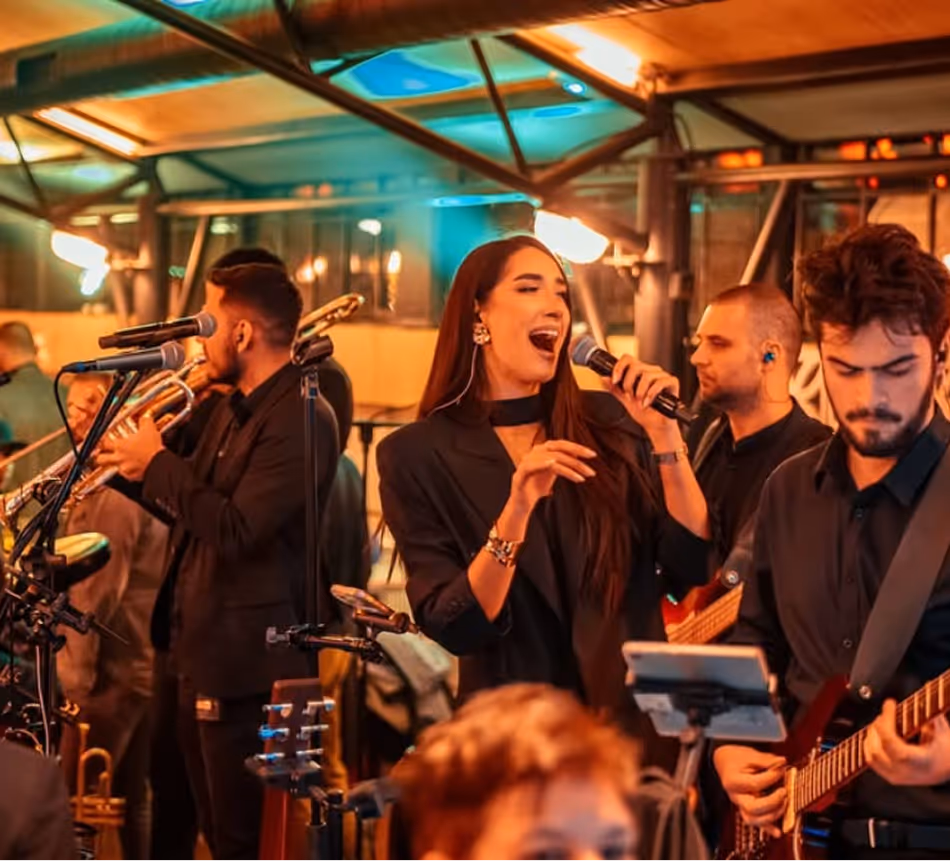 Muzičari na sceni: žena peva, muškarac svira gitaru i drugi sviraju trubu i bas.