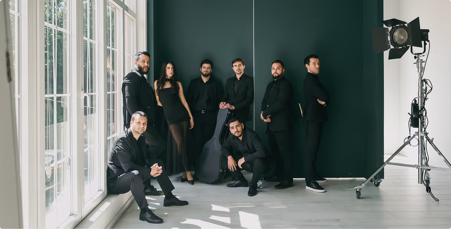Grupa ljudi u crnoj odeći pozira u svetloj sobi sa velikim prozorima i muzičkim koferom.