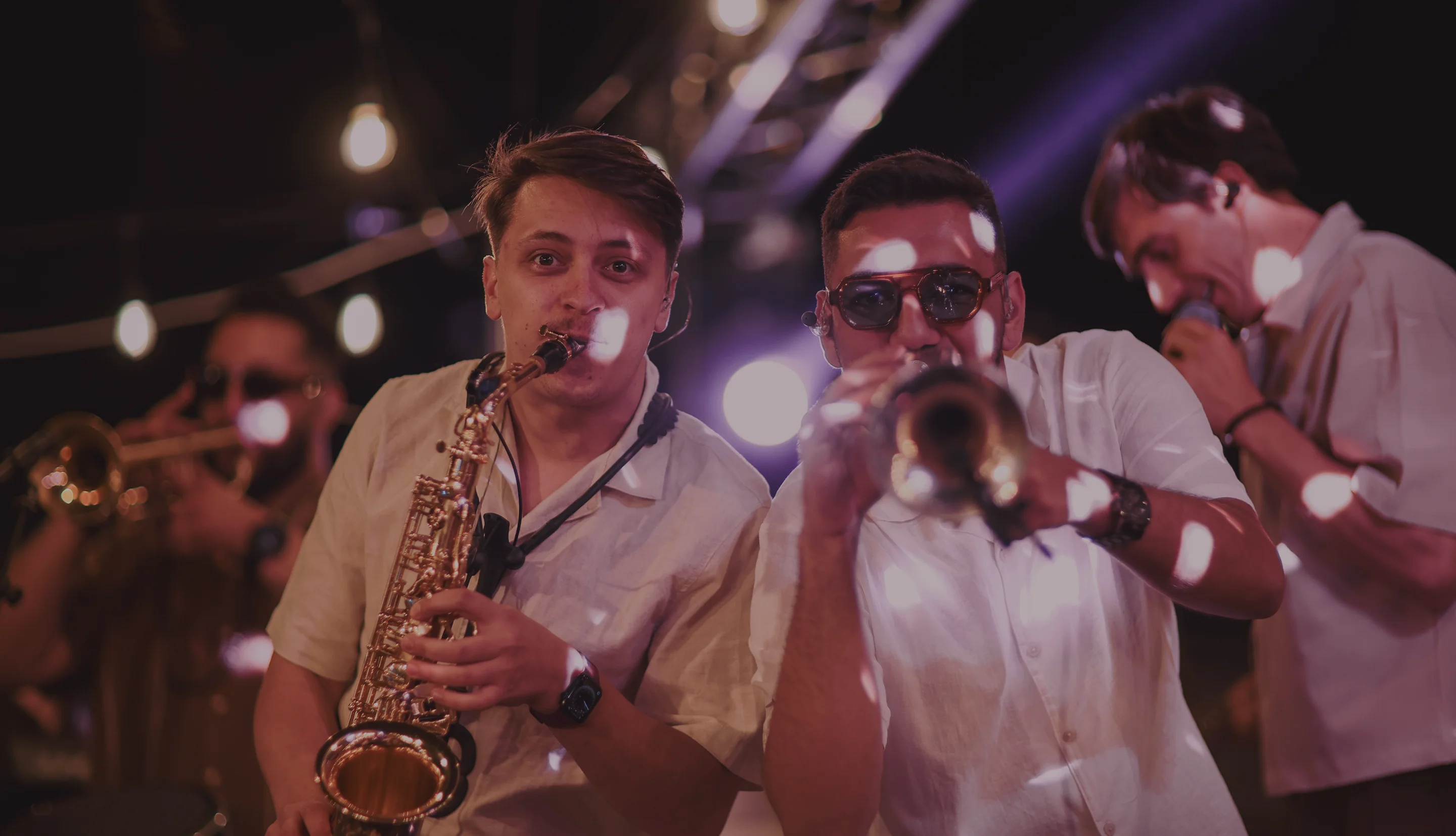 Četvorka muzičara svira saksofon, trubu i u košuljama na pozornici sa svetlima u pozadini.