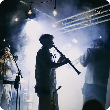 Tri osobe nastupaju sa duvačkim instrumentima na sceni punoj dima i svetala.