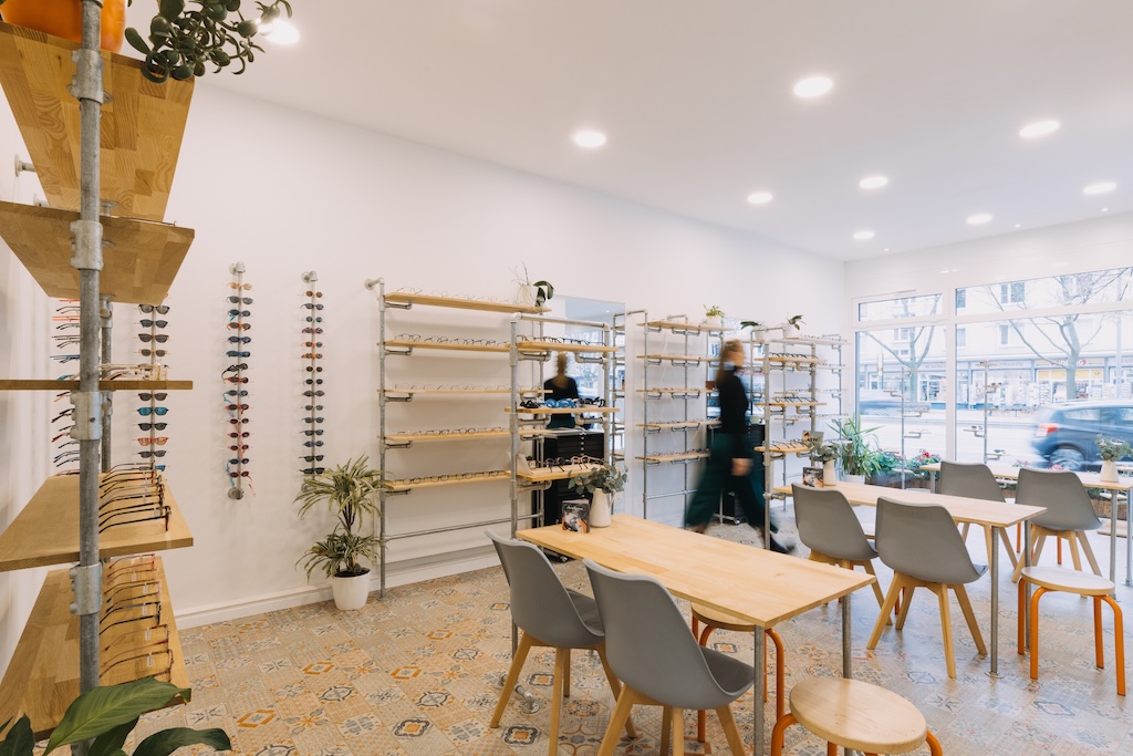 Helles Optikergeschäft mit moderner Brillenwand, Holztischen, Stühlen und großer Fensterfront.