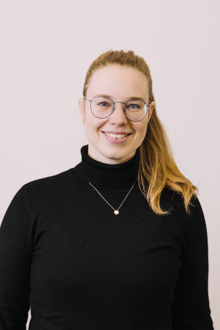 Frau mit hellbraunem Pferdeschwanz, Brille und schwarzem Rollkragenpullover lächelt vor hellem Hintergrund.