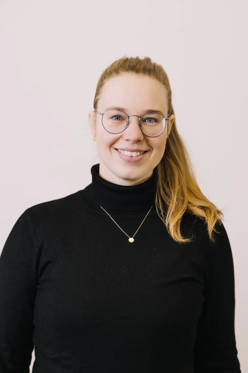 Frau mit hellbraunem Pferdeschwanz, Brille und schwarzem Rollkragenpullover lächelt vor hellem Hintergrund.
