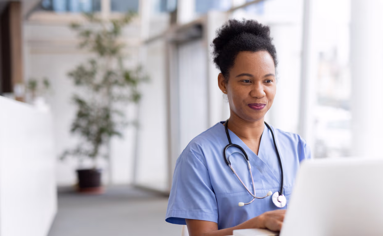 Smiling nurse in blue scrubs with stethoscope using a laptop in a bright indoor setting.