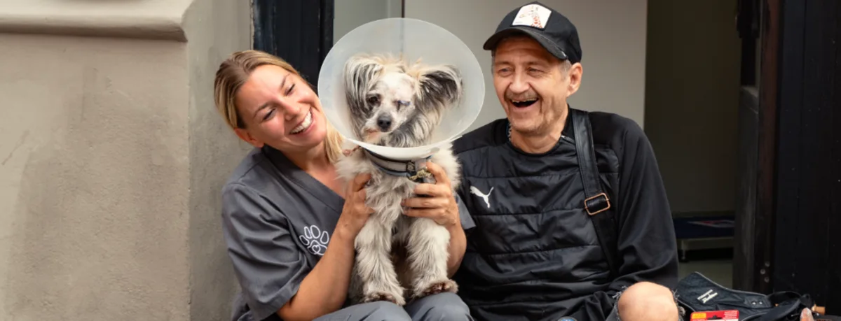 A smiling woman in veterinary scrubs holds a small dog wearing a protective cone, sitting next to a man in a black cap and jacket who is also smiling.