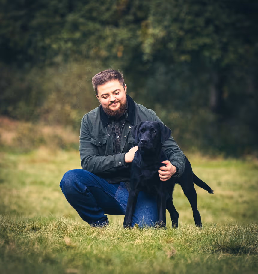 Man kneeling in grass outdoors with a black Labrador retriever dog beside him.