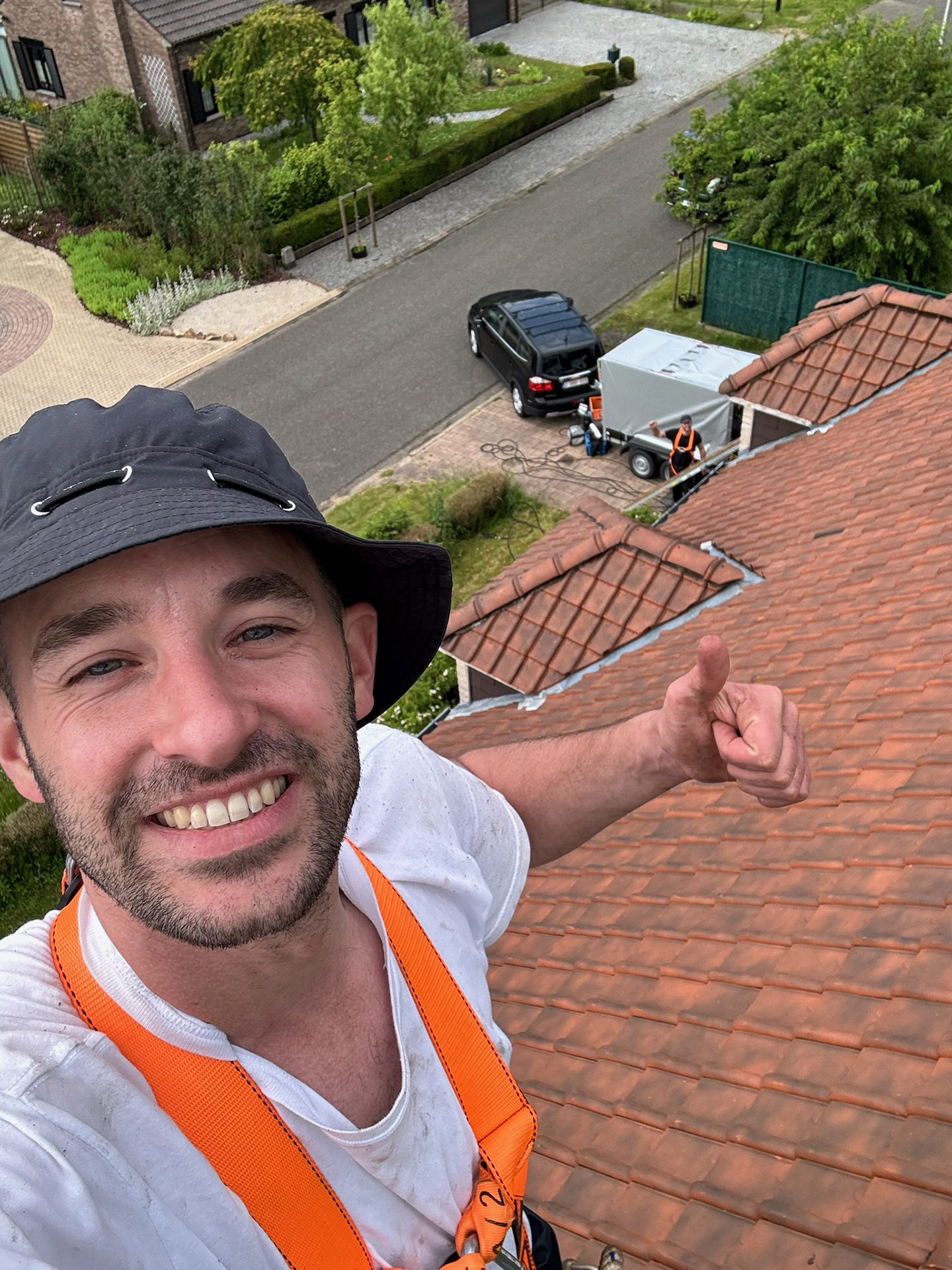 Mossmasters medewerker neemt selfie op pannendak met veiligheidsgordel tijdens dakreiniging met collega en aanhangwagen op straat