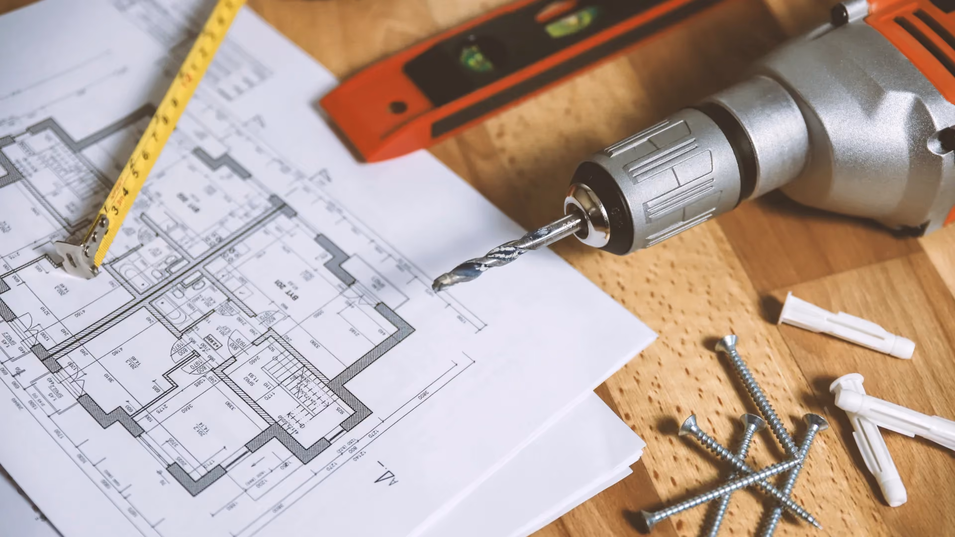 Bauplan mit Akkuschrauber, Wasserwaage, Maßband und Schrauben auf einem Holztisch