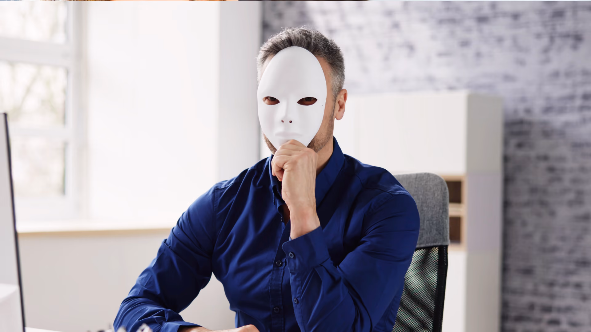 Mann am Schreibtisch versteckt sein Gesicht hinter einer Maske