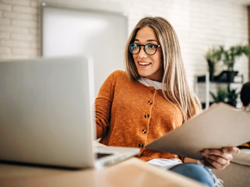 Frau mit Brille schaut auf den Bildschirm Ihres Laptops und hält ein Klemmbrett in der Hand