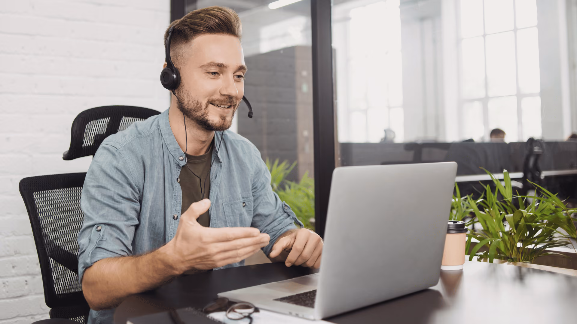 Mann mit Headset sitzt vor seinem Laptop und gestikuliert Richtung Bildschirm