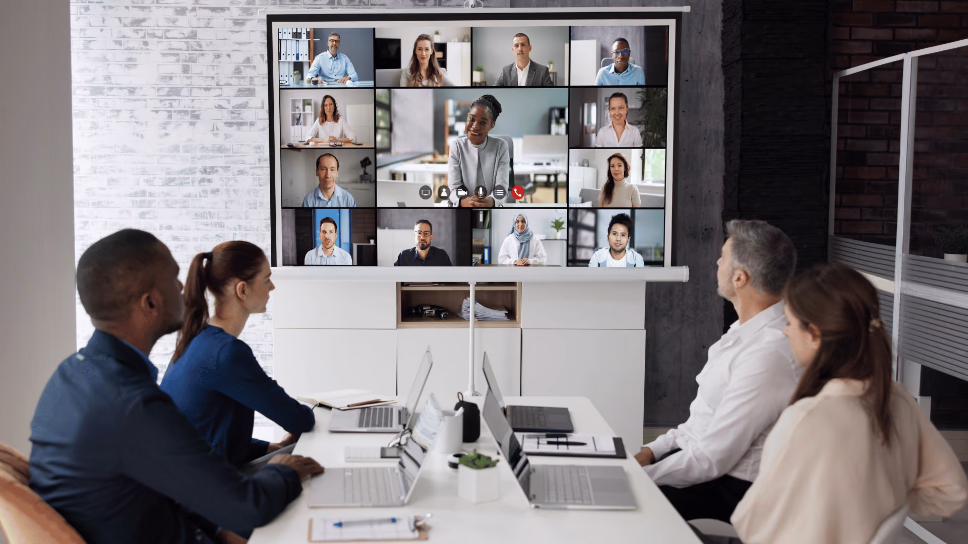 Vier Personen in einem Büroraum richten sich zu einer Leinwand auf der ein Online-Meeting übertragen wird