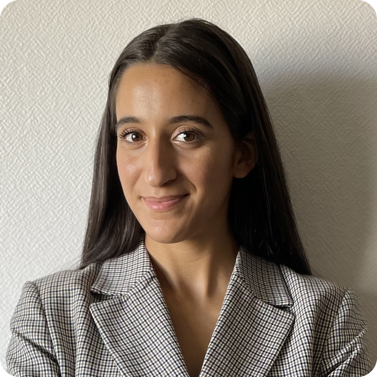 Femme souriante aux cheveux longs portant une veste à carreaux avant un mur blanc texturé.
