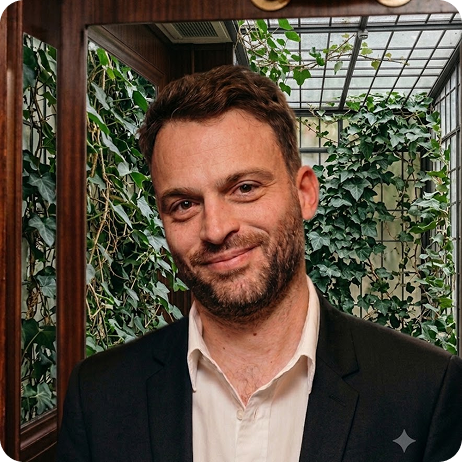 Homme souriant en costume devant une vitre avec des plantes vertes en arrière-plan.