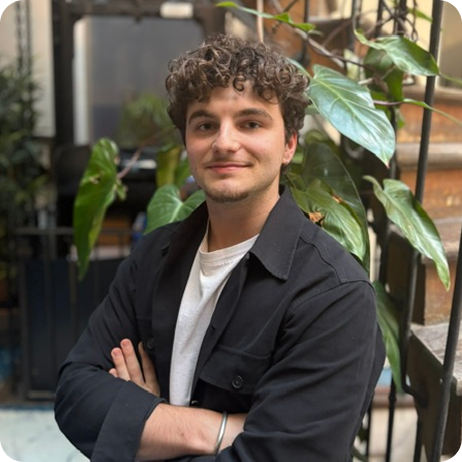 Un homme souriant portant un costume noir et une chemise blanche devant un mur de plantes vertes.