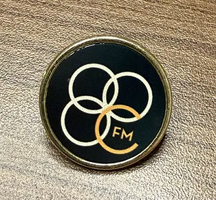 Round black pin with four connected gold rings and letters 'FM' on one ring.