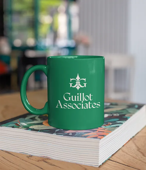 Green mug with Guillot Associates logo on a colorful book on a wooden table indoors.