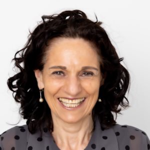 Smiling woman with curly dark hair wearing pearl earrings and a gray polka dot blouse.