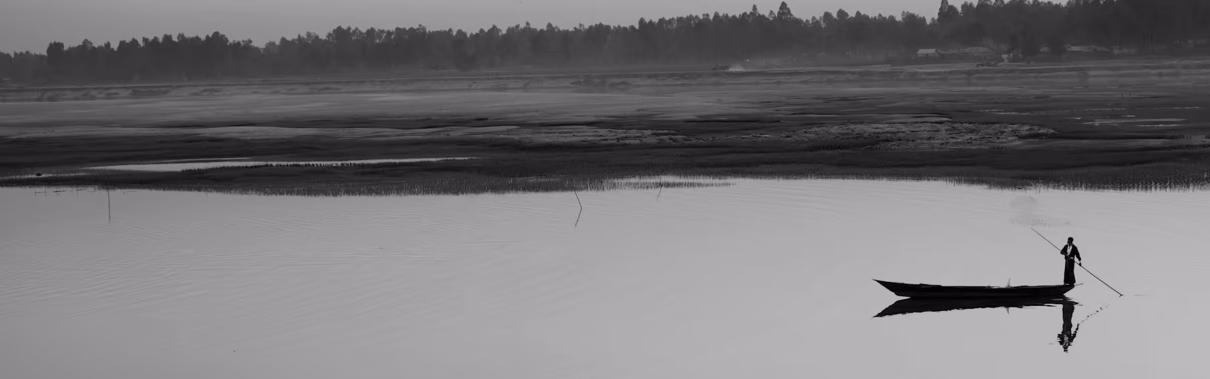 Black and white image of a person standing on a narrow boat on calm water near a shore with trees.