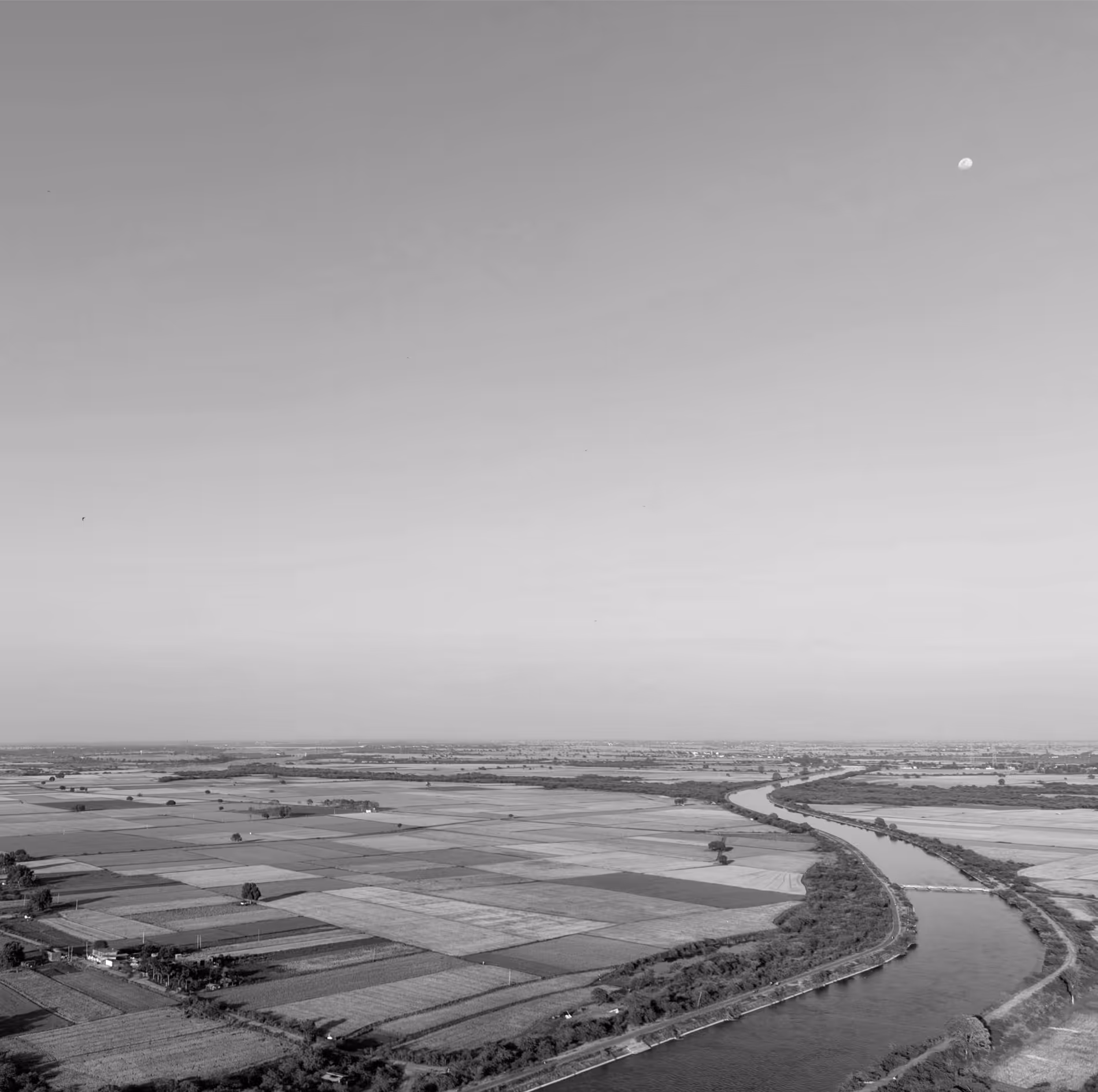Aerial view of patchwork farmland with a winding river under a clear sky with a visible moon.