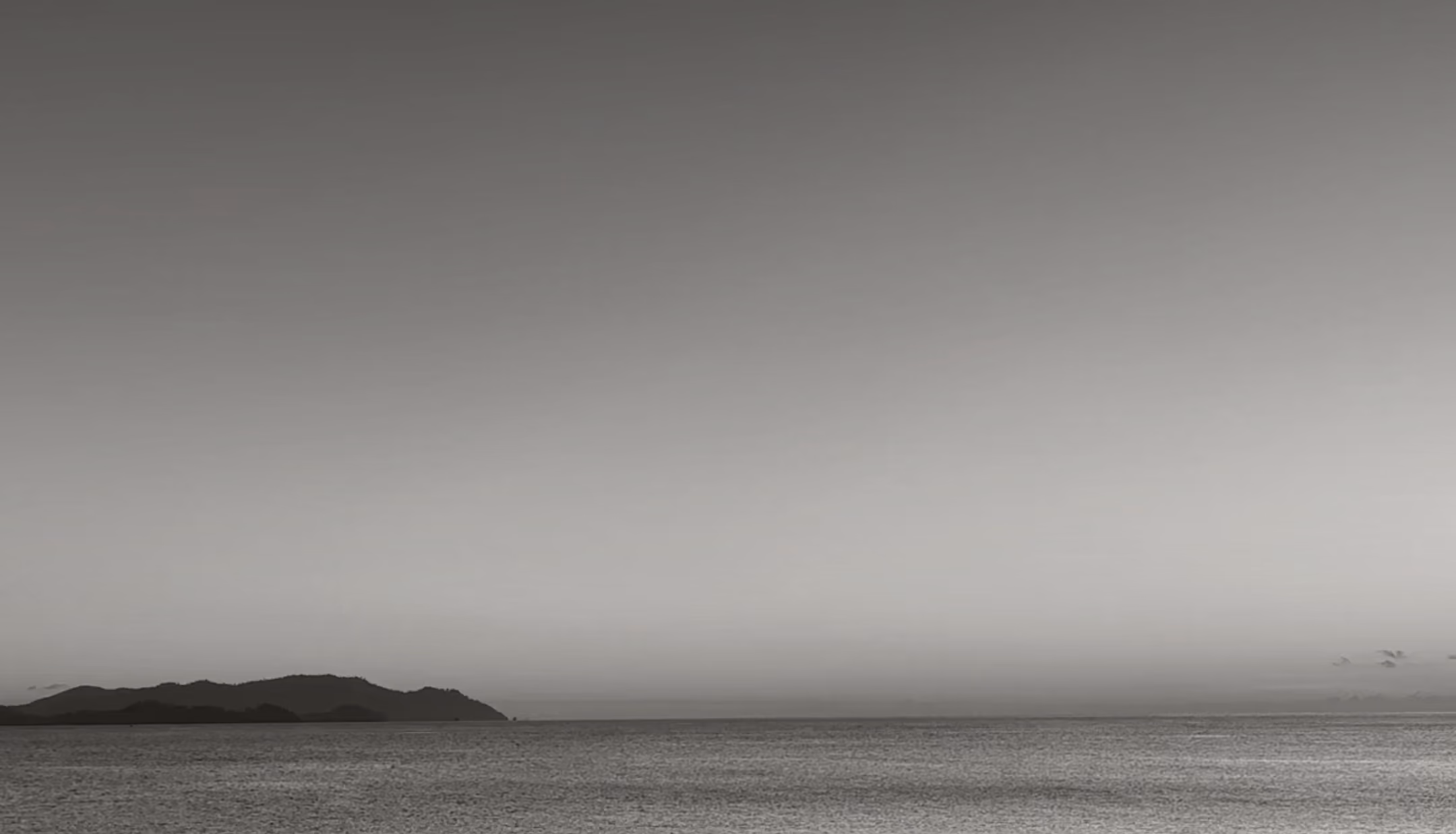 Black and white photo of calm sea with a low mountain range in the distance under clear sky.