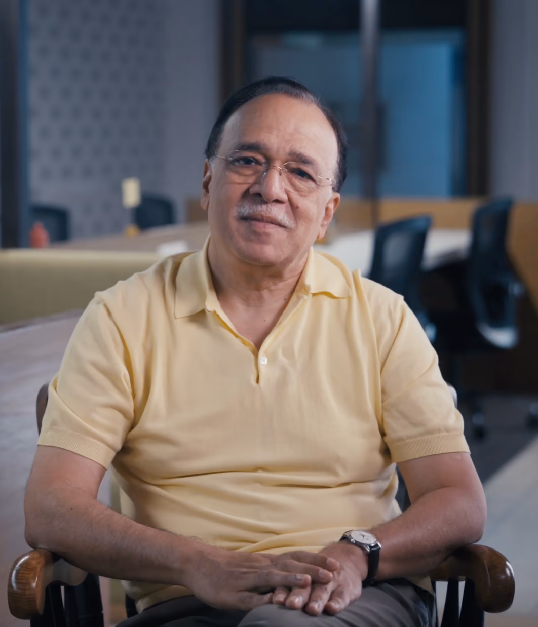 Older man with glasses and mustache sitting in chair wearing a yellow polo shirt in an office.