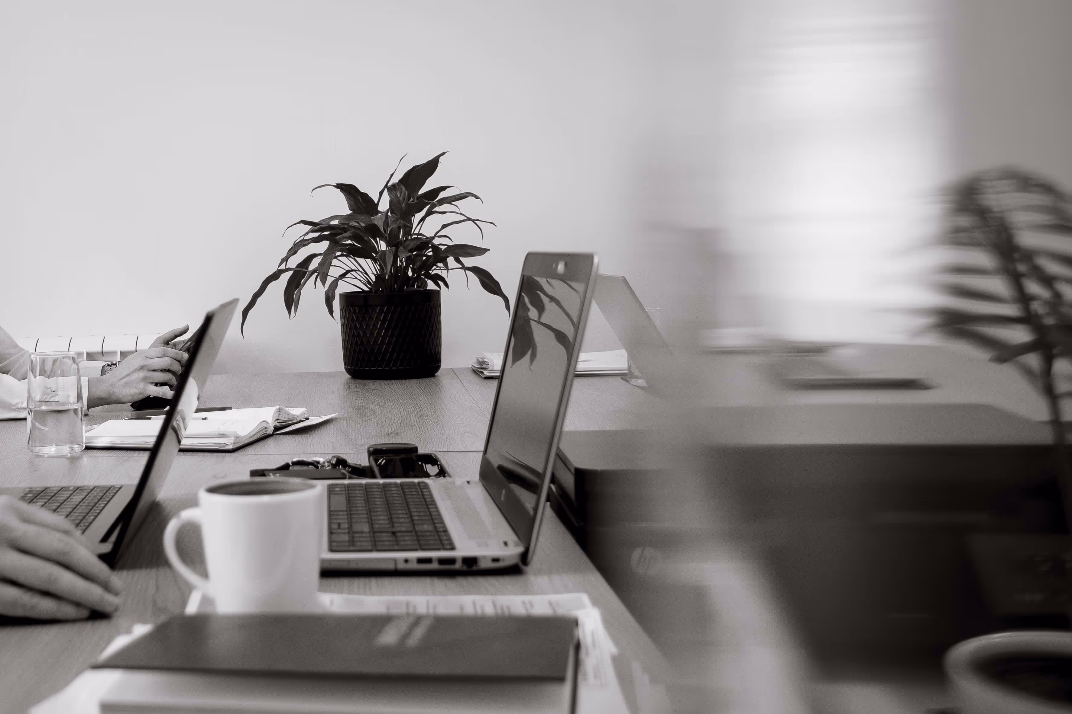 Office desk with laptops, a coffee mug, notebooks, a plant, and blurred motion of a person.