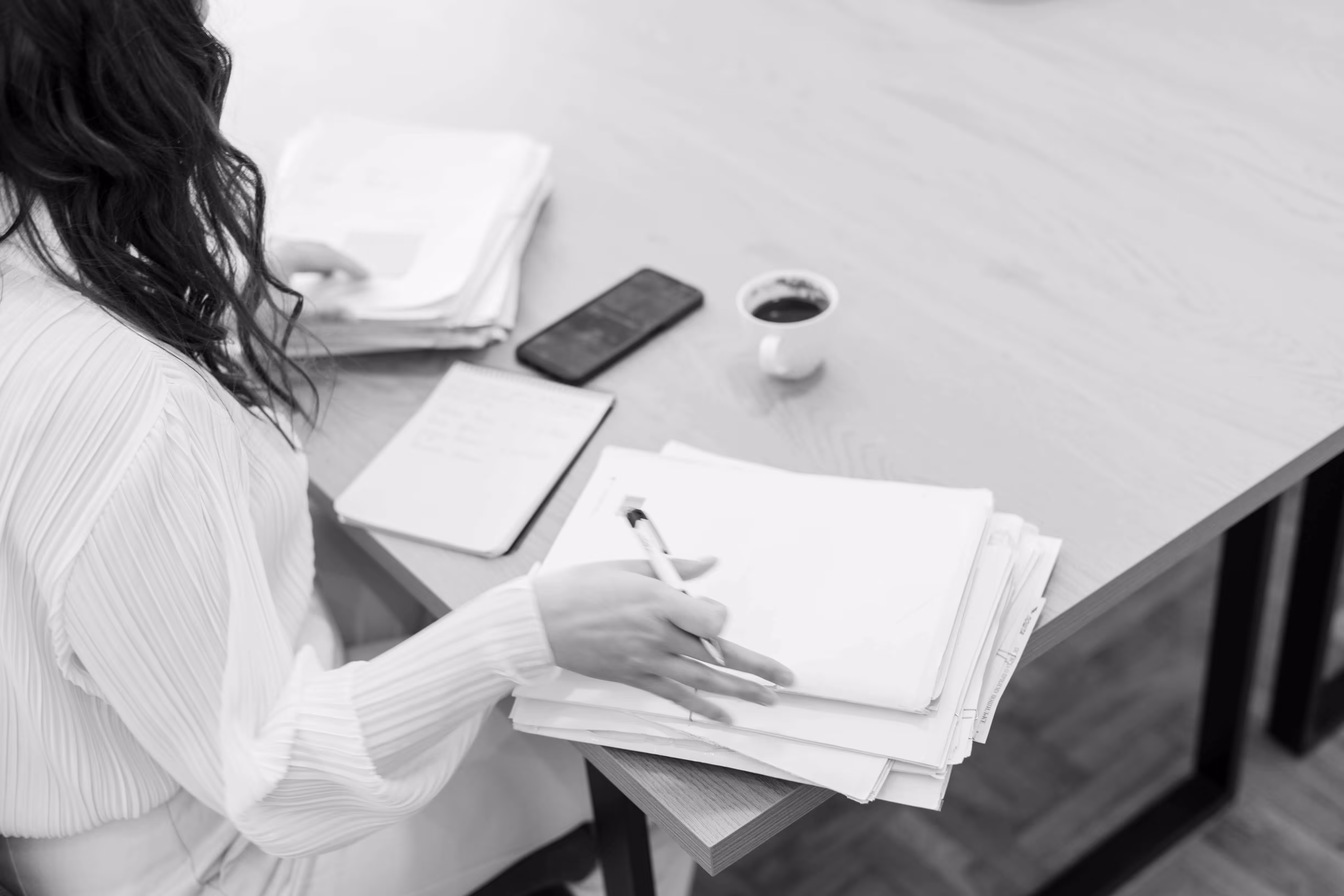 Person organizing documents on a desk with a notebook, smartphone, and coffee cup nearby.