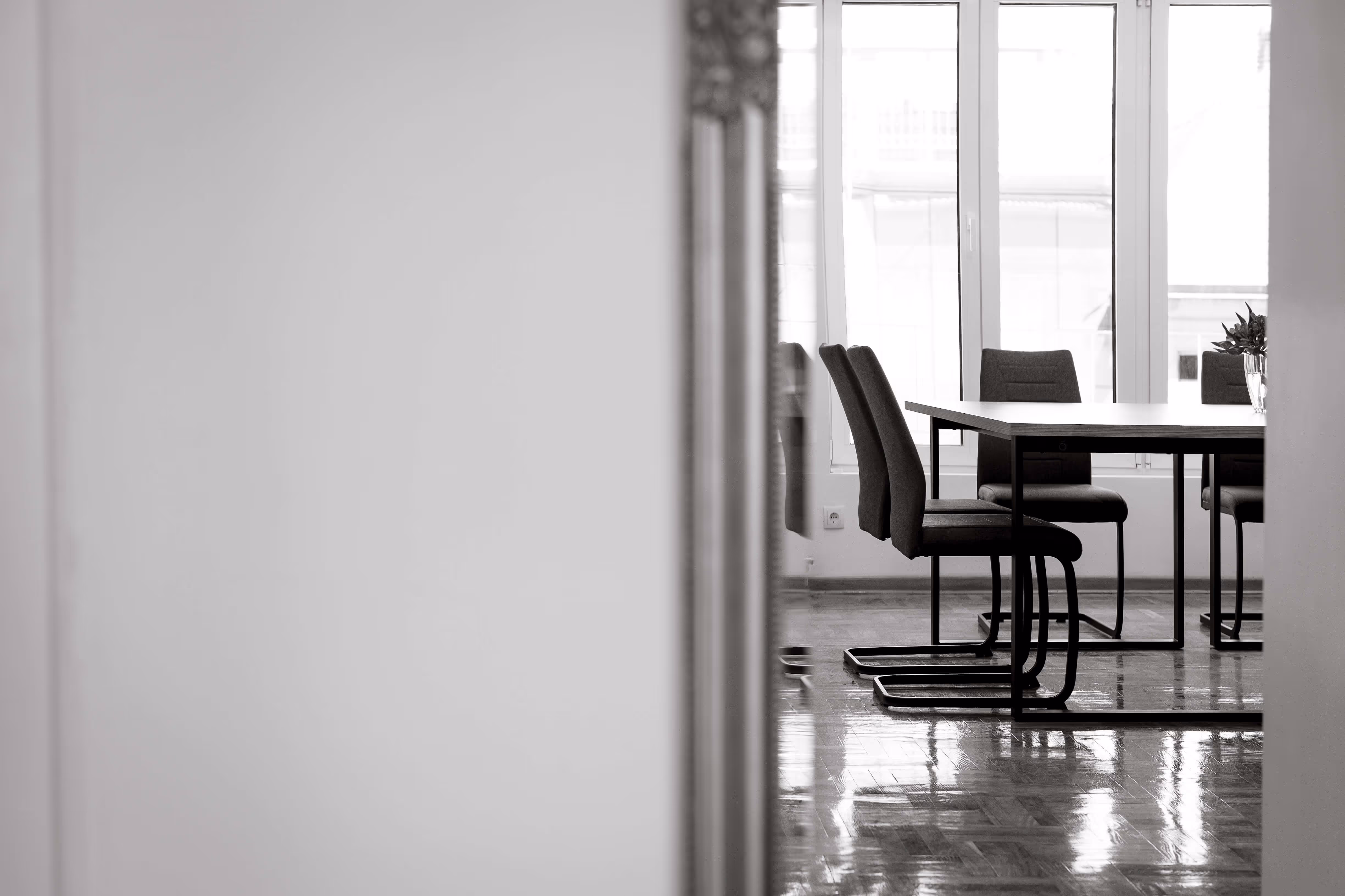 Black and white photo of a dining table with chairs and a vase with flowers near large windows.