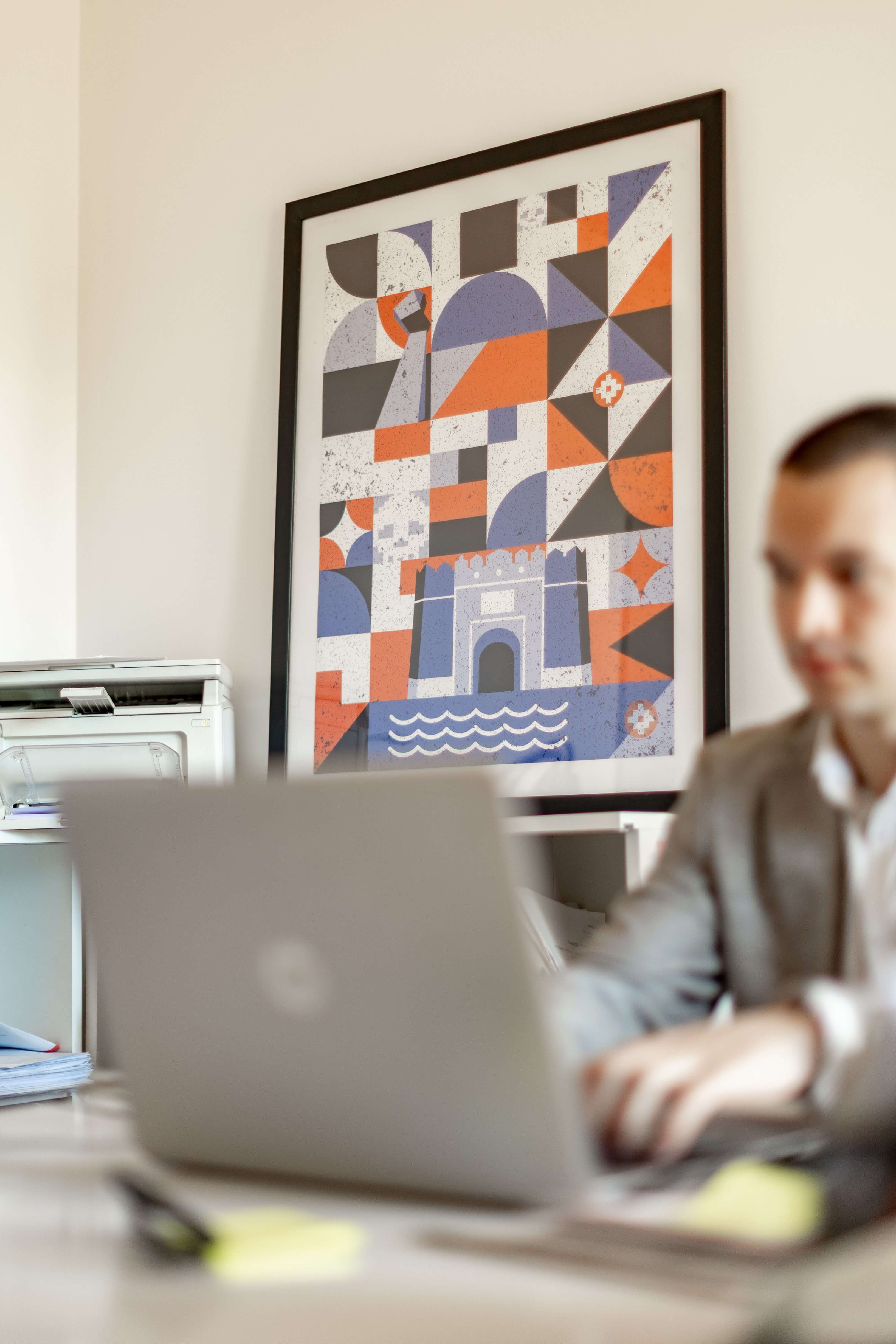 Abstract geometric art in shades of orange, purple, and black framed on an office wall behind a laptop.