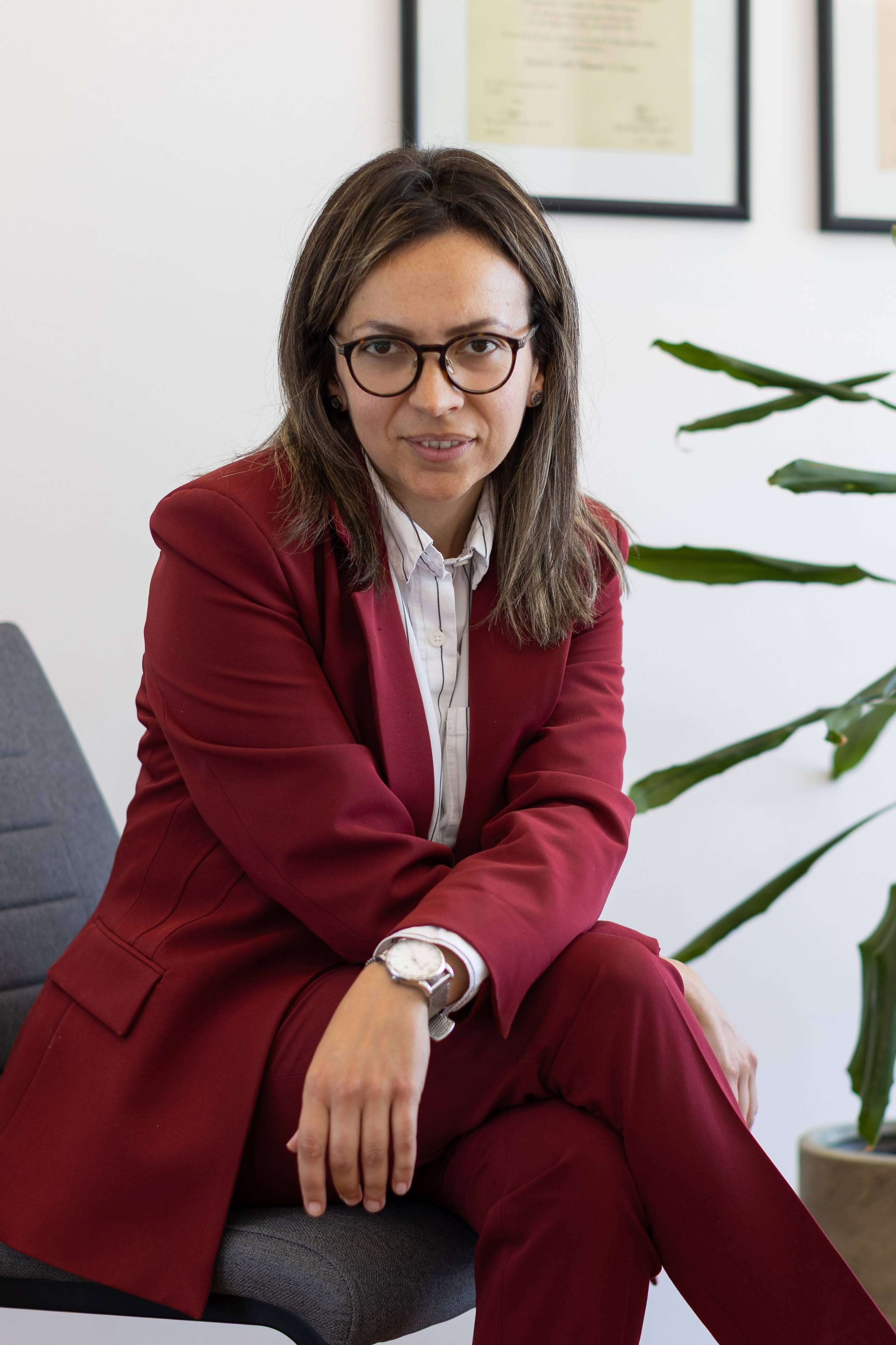 Woman in glasses wearing a red suit seated on a gray chair in an office with framed certificates.