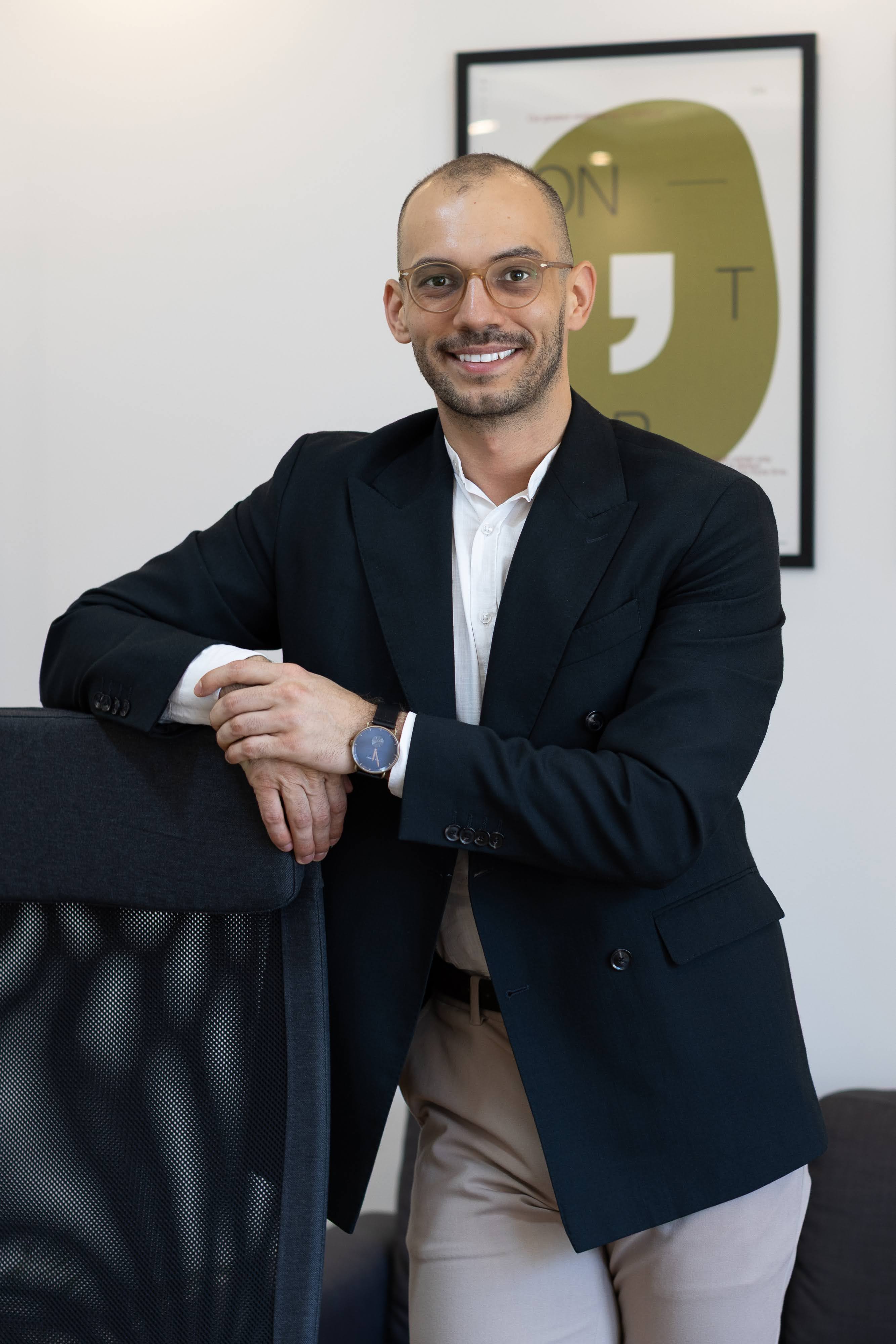 Smiling man in glasses wearing black blazer leaning on a black office chair in a modern office.