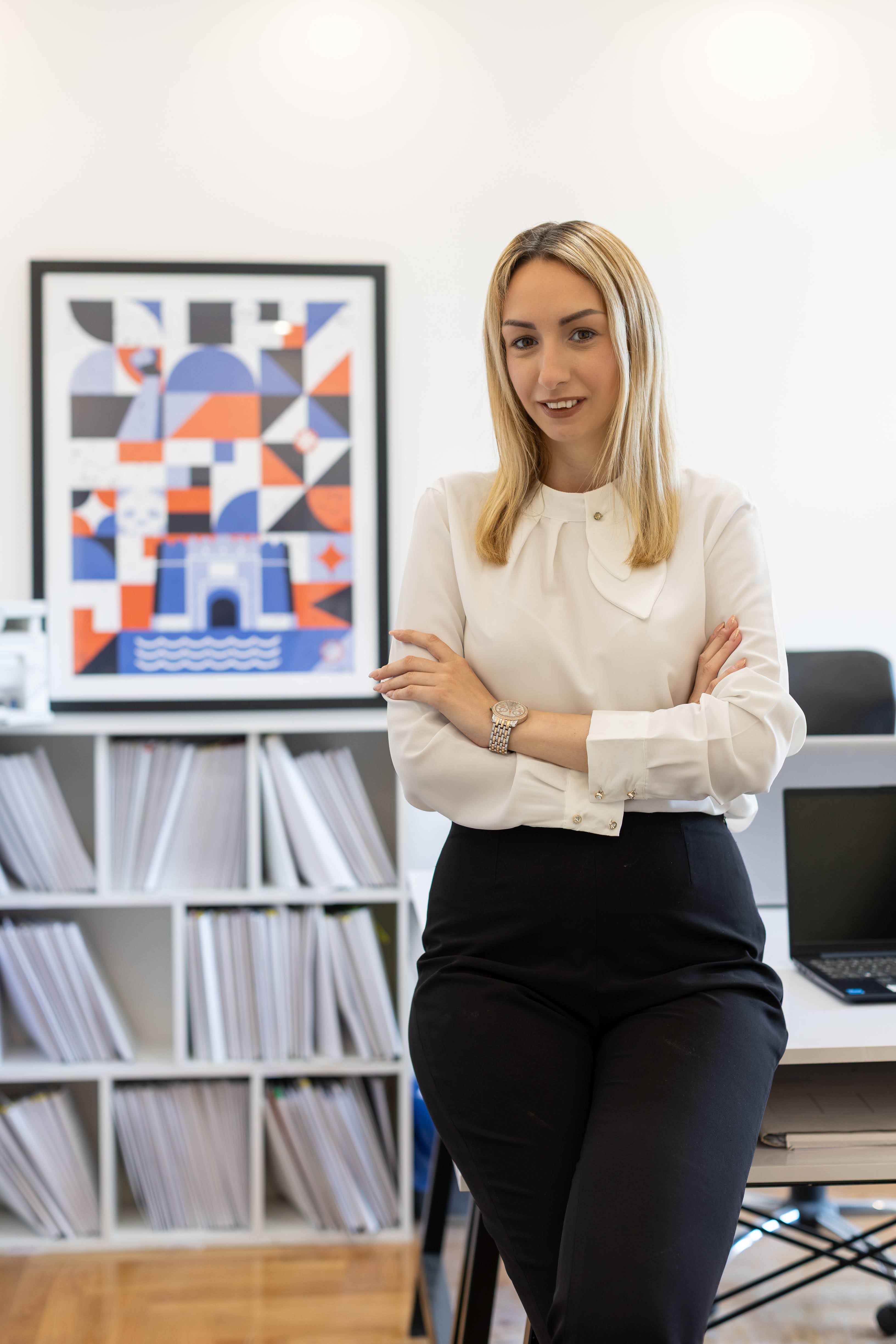 Woman in white blouse and black pants standing with arms crossed in modern office space.