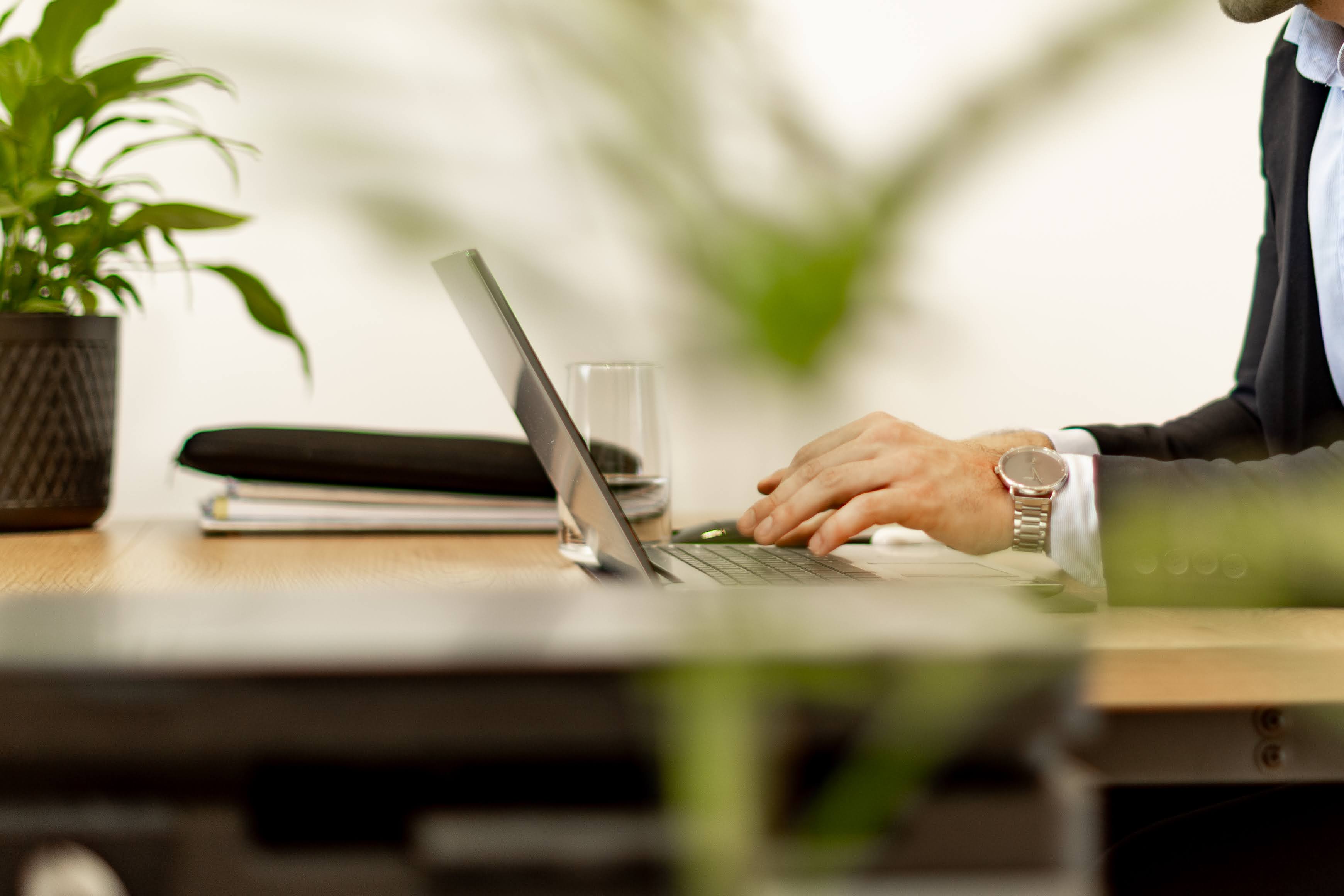 Person in a suit typing on a laptop at a desk with plants, folders, and a glass of water nearby.