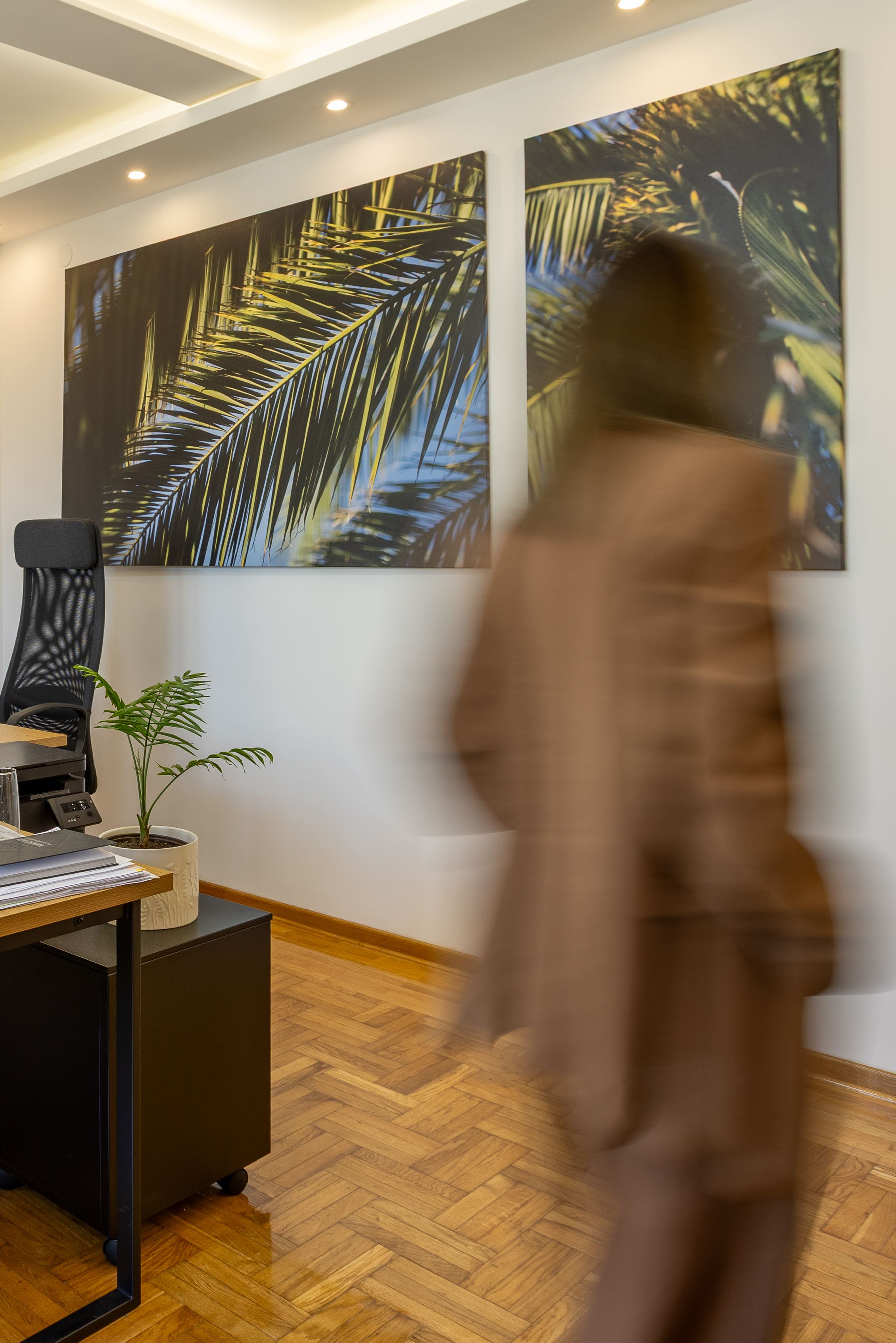 Blurred person walking in office with wooden floor, desk, chair, plant, and palm leaf wall art.