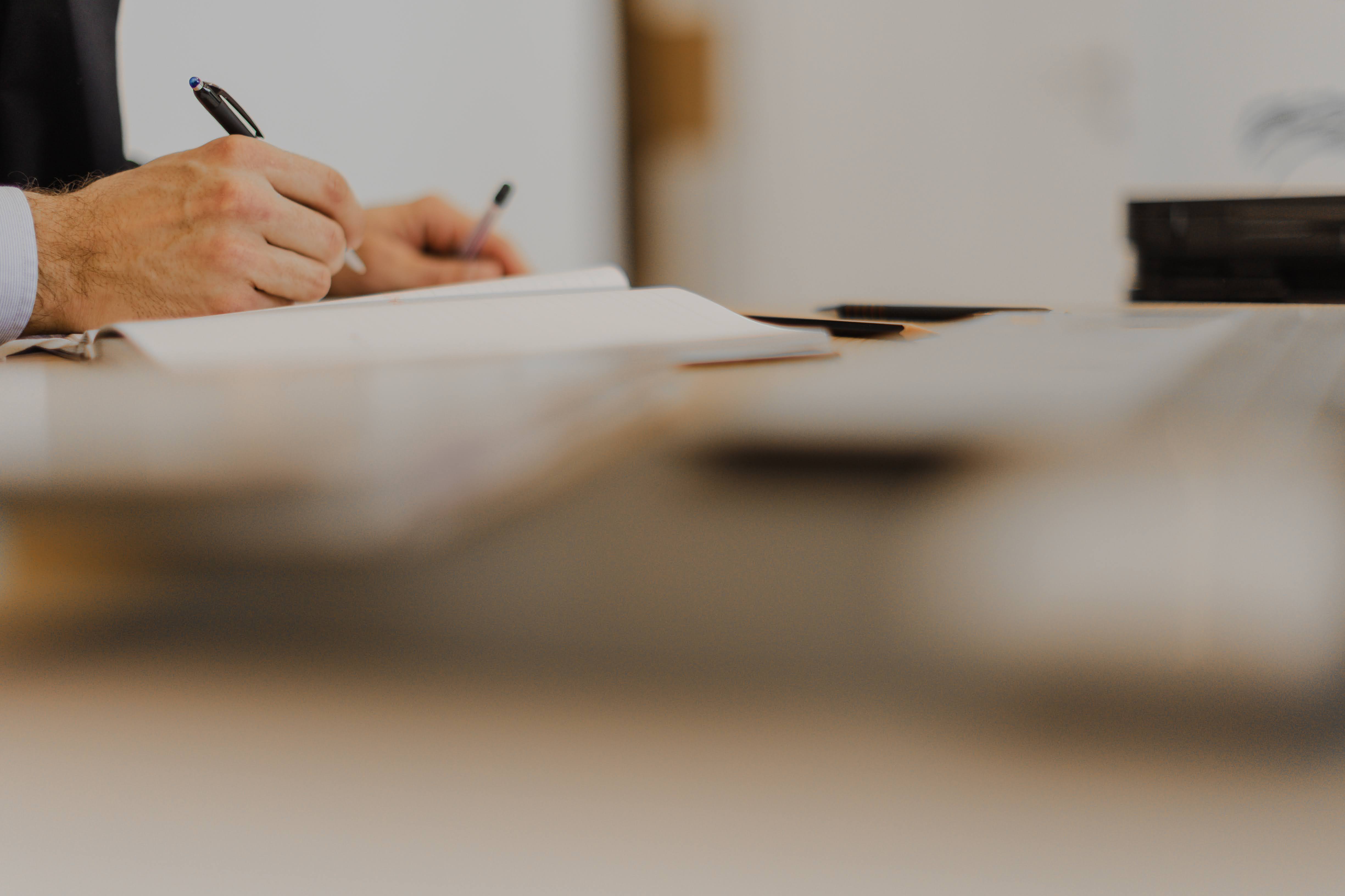 Person writing with pen on paper at desk with blurred laptop and notebook nearby.
