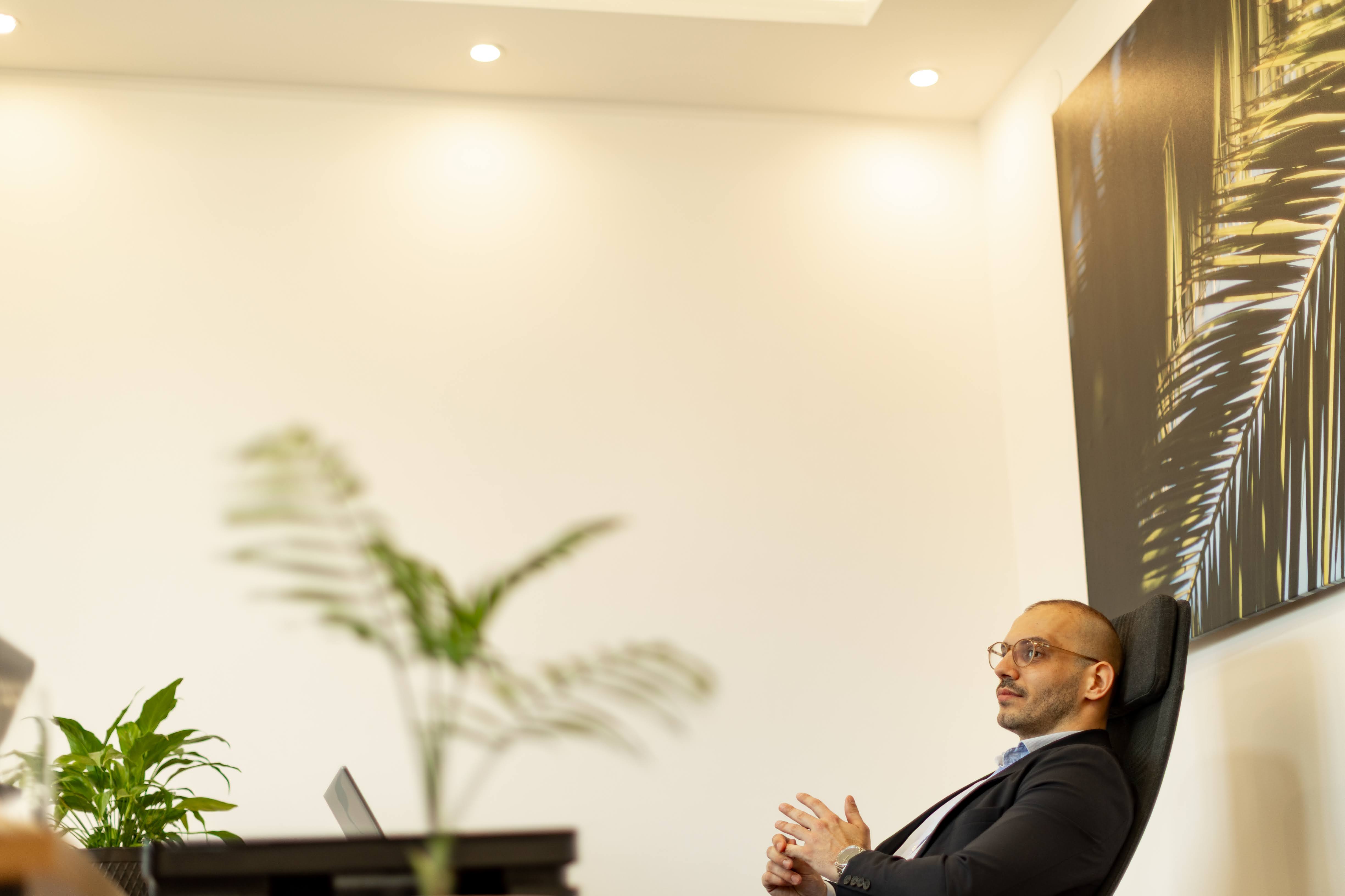 Man in glasses and suit relaxing in an office chair by a desk with plants and laptop.