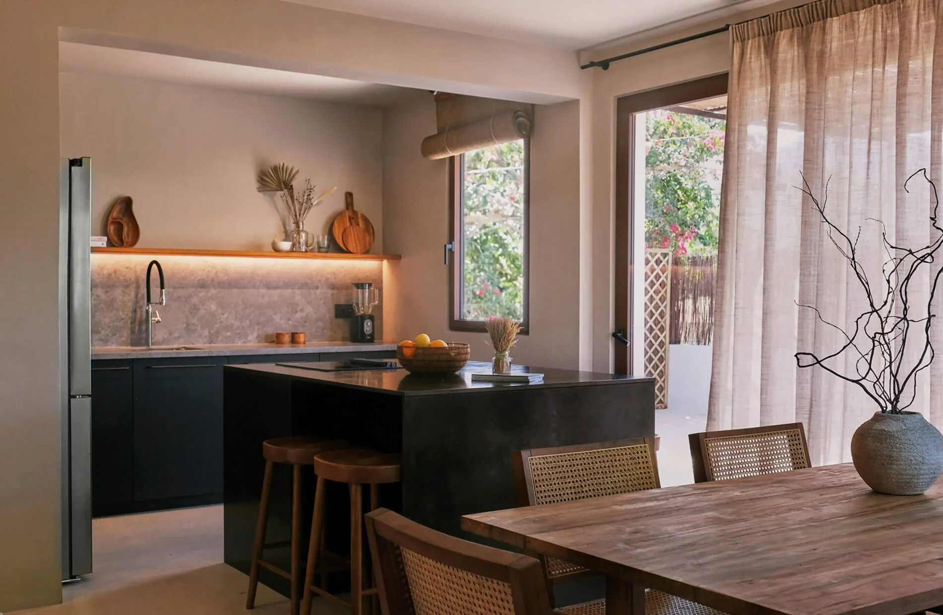 Open-plan kitchen and dining area design at Sa Caleta, Ibiza, highlighting a matte black island, wooden bar stools, and a rustic wooden dining table.