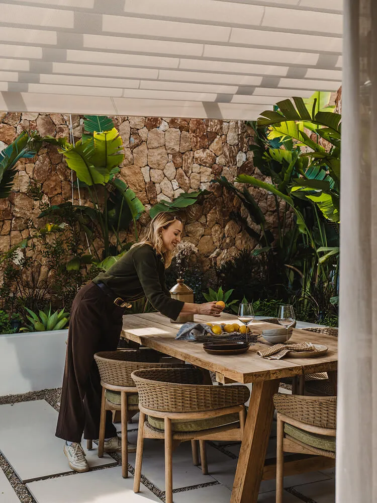 Mujer arreglando limones sobre una mesa de madera en un acogedor comedor al aire libre con paredes de piedra y plantas.