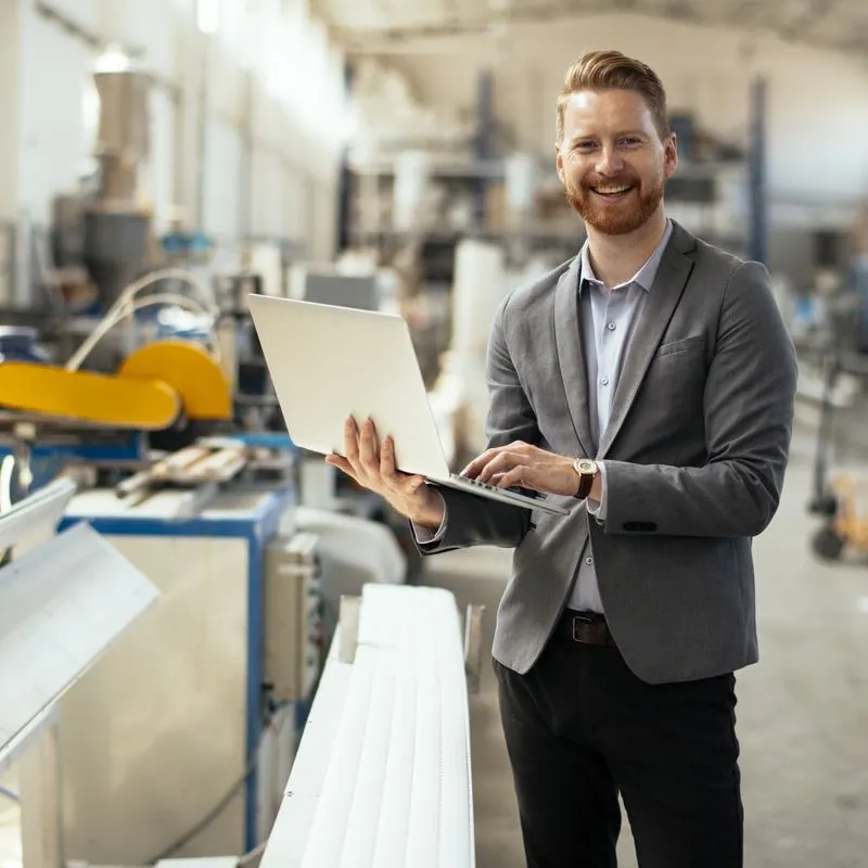 Smiling man with red hair and beard wearing a gray blazer and holding a laptop in an industrial manufacturing facility.