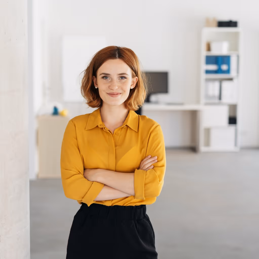 Confident woman with short brown hair in a mustard yellow button-up shirt and black pants standing with arms crossed in a bright office setting.