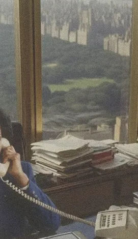 Person on telephone with window showing city park and buildings, desk stacked with papers nearby.