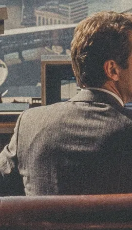 Man with brown hair in a gray suit sitting indoors facing away near a window.