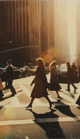 People crossing a city street at a crosswalk with sunlight casting long shadows on the pavement.