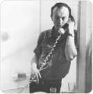Black and white photo of a man holding a corded phone to his ear, speaking.