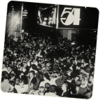Black and white photo of a large crowd at a night event with posters in the background.