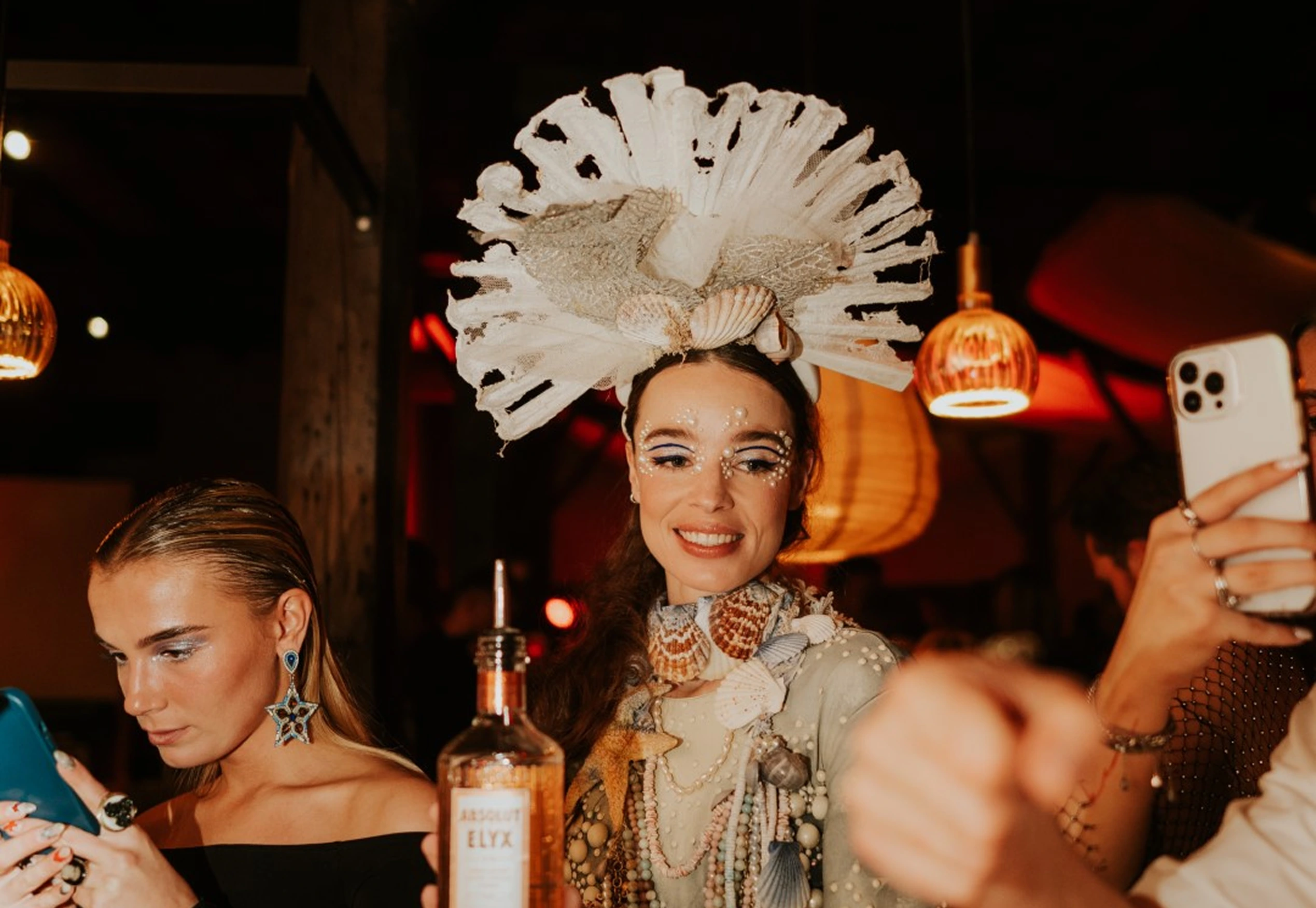 Performer in a shell headdress surrounded by guests raising their phones at a themed party