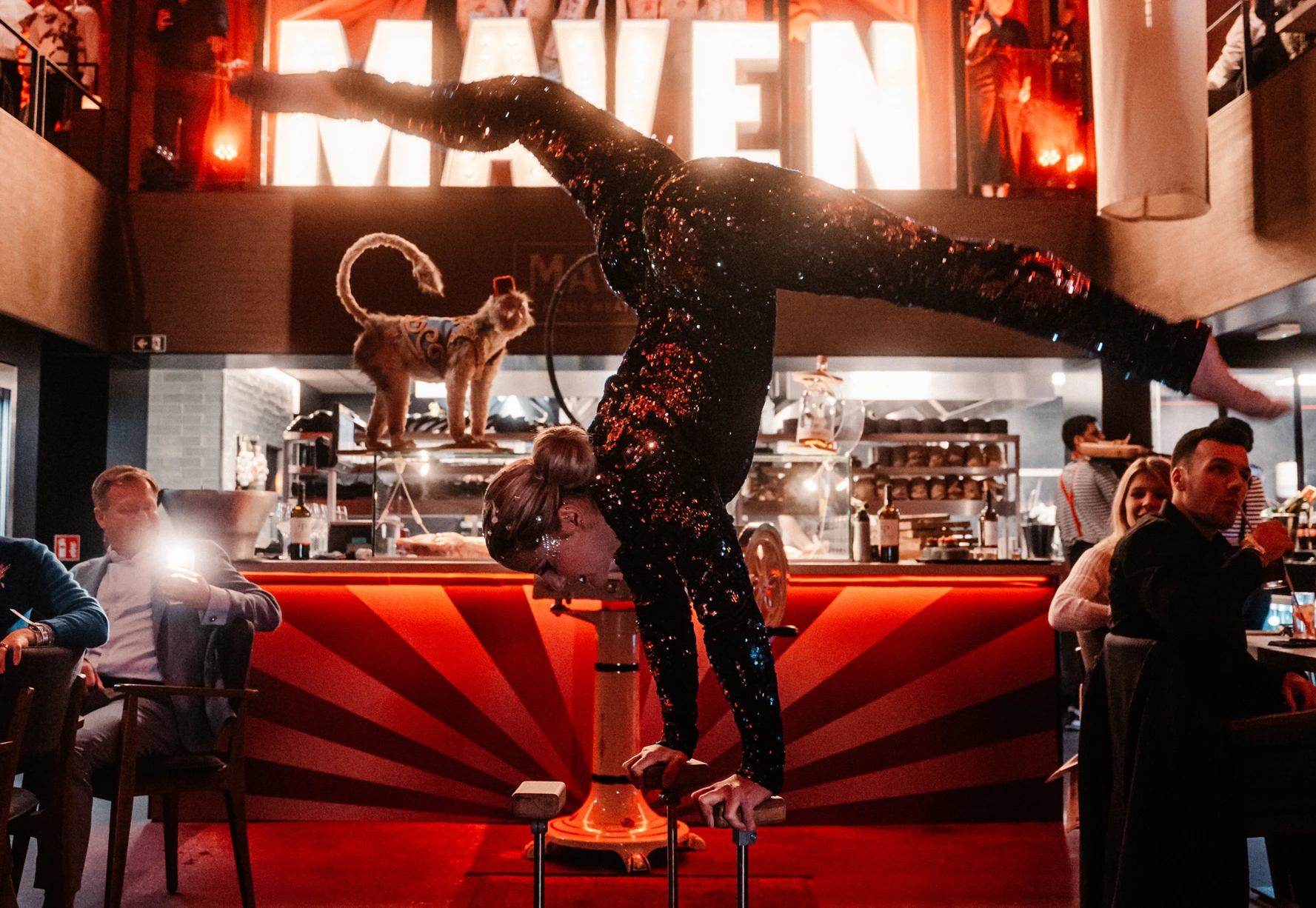 Acrobat holding a handstand in front of the illuminated MAVEN sign during an evening at the bar